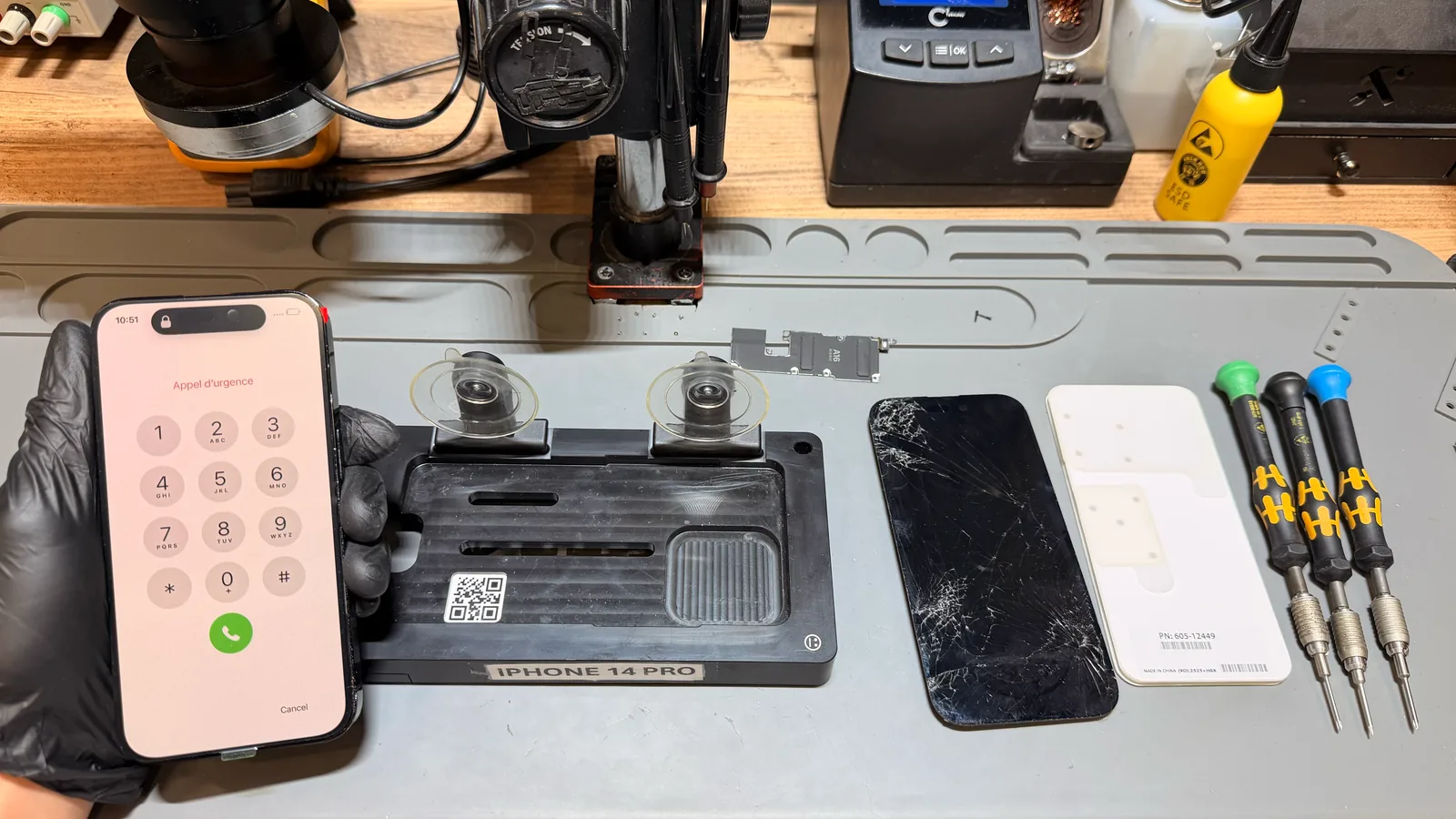 Siyah eldivenli el, ekranı yenilenip açılan bir iPhone 14 Pro'yu tutuyor (acil arama ekranı açık); yanında ekran ayırma aparatı, kırık eski ekran ve orijinal parça ambalajı.