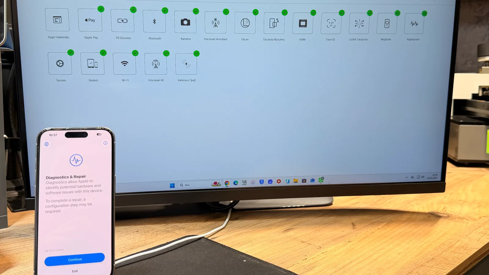 Atölye monitöründe Apple onarım sonrası tanılama ekranı; tüm bileşenler yeşil onay işaretli. Önde 'Diagnostics & Repair' ekranı açık bir iPhone.