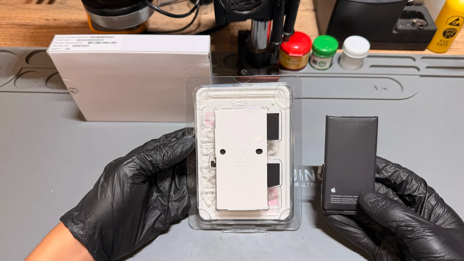 Siyah eldivenli eller iki iPhone pilini yan yana tutuyor: solda ambalajındaki yeni orijinal pil, sağda çıkarılan eski pil; arkada orijinal parça kutusu ve ESD çalışma matı.